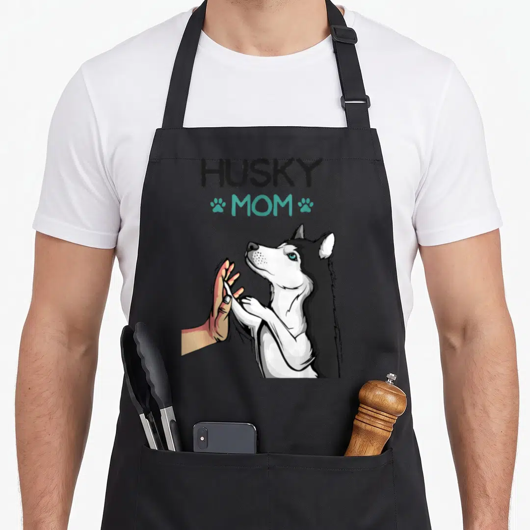 Mummy dog apron - Cartoon husky high-fiving human hand with HUSKY MOM text and teal paw prints — shown on a black cotton canvas apron against a white background