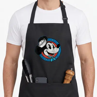 Cruise distressed apron - Cartoon Mickey Mouse in sailor hat and bandana inside a blue circular porthole frame — shown on a black cotton canvas apron against a white background