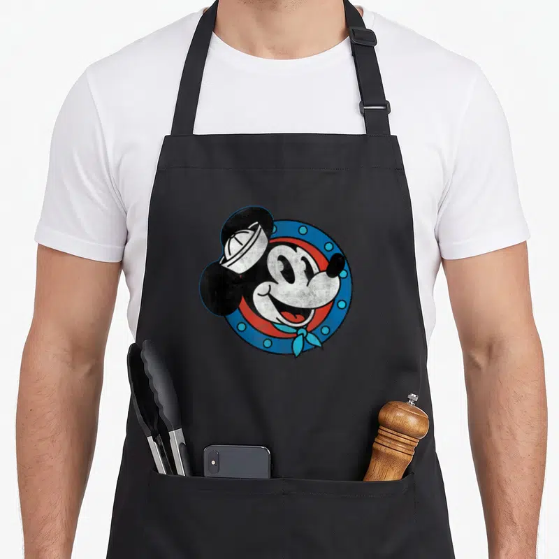 Cruise distressed apron - Cartoon Mickey Mouse in sailor hat and bandana inside a blue circular porthole frame — shown on a black cotton canvas apron against a white background