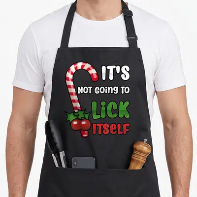 Itself candy cane apron - Cartoon candy cane with holly and text IT'S NOT GOING TO LICK ITSELF — shown on a black cotton canvas apron against a white background