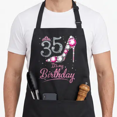 Born day queen apron - Diamond 35 high heel with pink crown and It's my Birthday text in glitter script — shown on a black cotton canvas apron against a white background