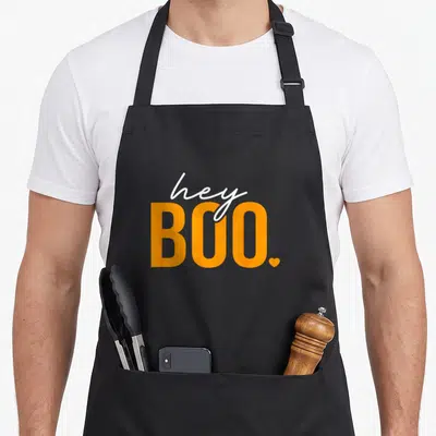 Hey halloween apron - Orange BOO text with white hey script and small heart punctuation — shown on a black cotton canvas apron against a white background