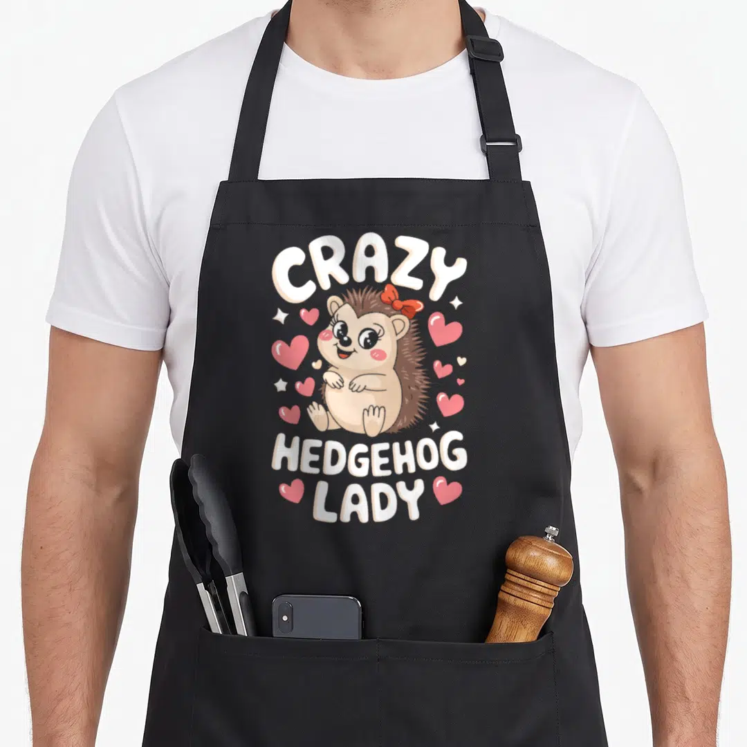 Crazy hedgehog queen apron - Cartoon tan hedgehog with red bow, pink hearts, and white text CRAZY HEDGEHOG LADY — shown on a black cotton canvas apron against a white background