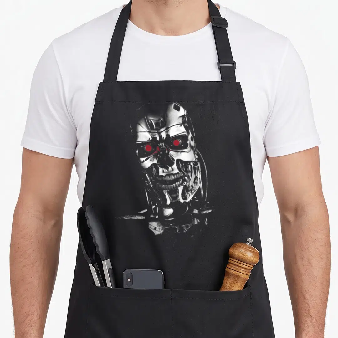 Terminator machine apron - Grey and white robotic skull with glowing red eyes on black background — shown on a black cotton canvas apron against a white background