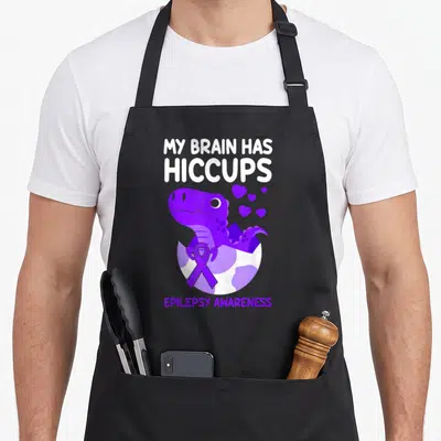 Hiccup ribbon apron - Purple dinosaur hatching from egg with ribbon and hearts under MY BRAIN HAS HICCUPS text — shown on a black cotton canvas apron against a white background