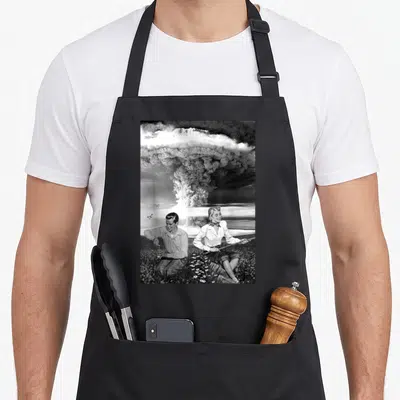 Atomic military apron - Mushroom cloud over couple sitting in flowers, grey white black photo style — shown on a black cotton canvas apron against a white background