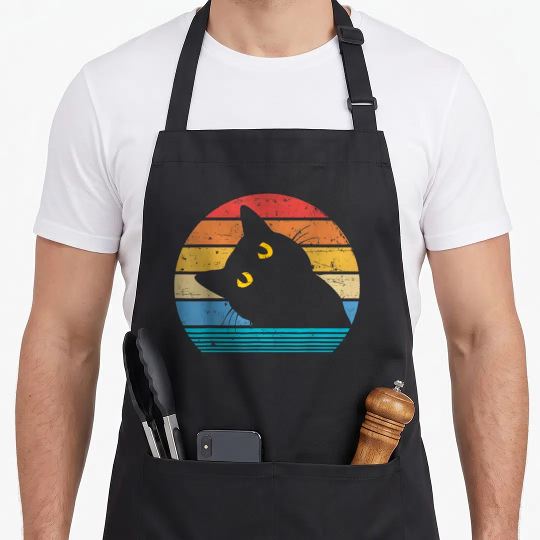 Kitty black cat apron - Grey cat silhouette over red orange yellow blue striped retro sun graphic — shown on a black cotton canvas apron against a white background