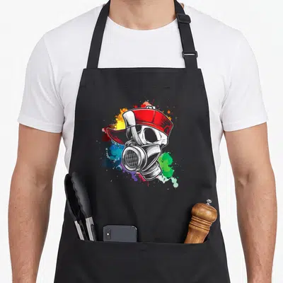 Gas tag apron - Cartoon skull in red cap and gas mask over multicolored paint splatter — shown on a black cotton canvas apron against a white background