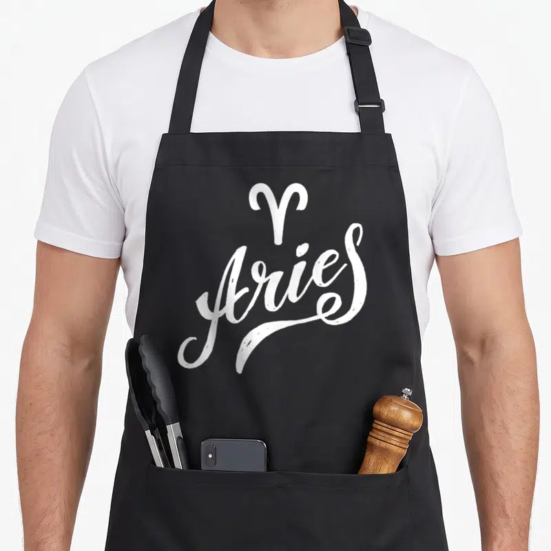 Funny zodiac aries kitchen apron - White Aries zodiac symbol and script text on dark background — shown on a black cotton canvas apron against a white background