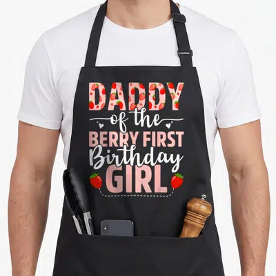 Strawberry daddy apron - Stacked text Daddy Berry First Birthday Girl filled with pink cartoon strawberries and dotted — shown on a black cotton canvas apron against a white background