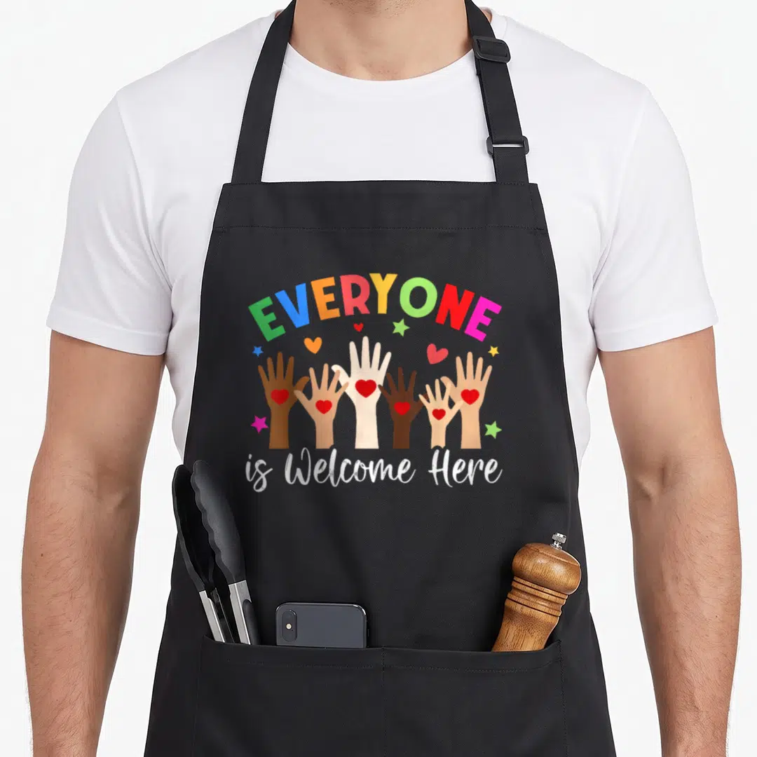 Idaho teacher apron - Rainbow text above six diverse hands holding red hearts with stars and script reading is Welcome — shown on a black cotton canvas apron against a white background