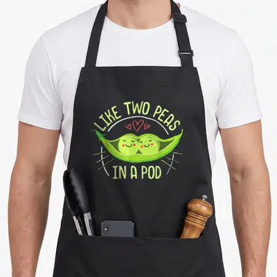 Pea pod aprons - Cartoon green pea pod with two smiling peas and three red hearts under arching text — shown on a black cotton canvas apron against a white background