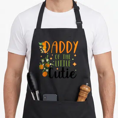 Cutie daddy apron - Daddy of the Little Cutie text with clementines and sparkles — shown on a black cotton canvas apron against a white background