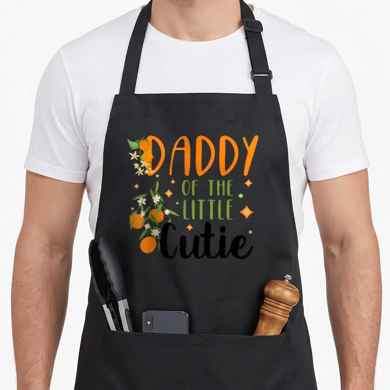 Cutie daddy apron - Daddy of the Little Cutie text with clementines and sparkles — shown on a black cotton canvas apron against a white background