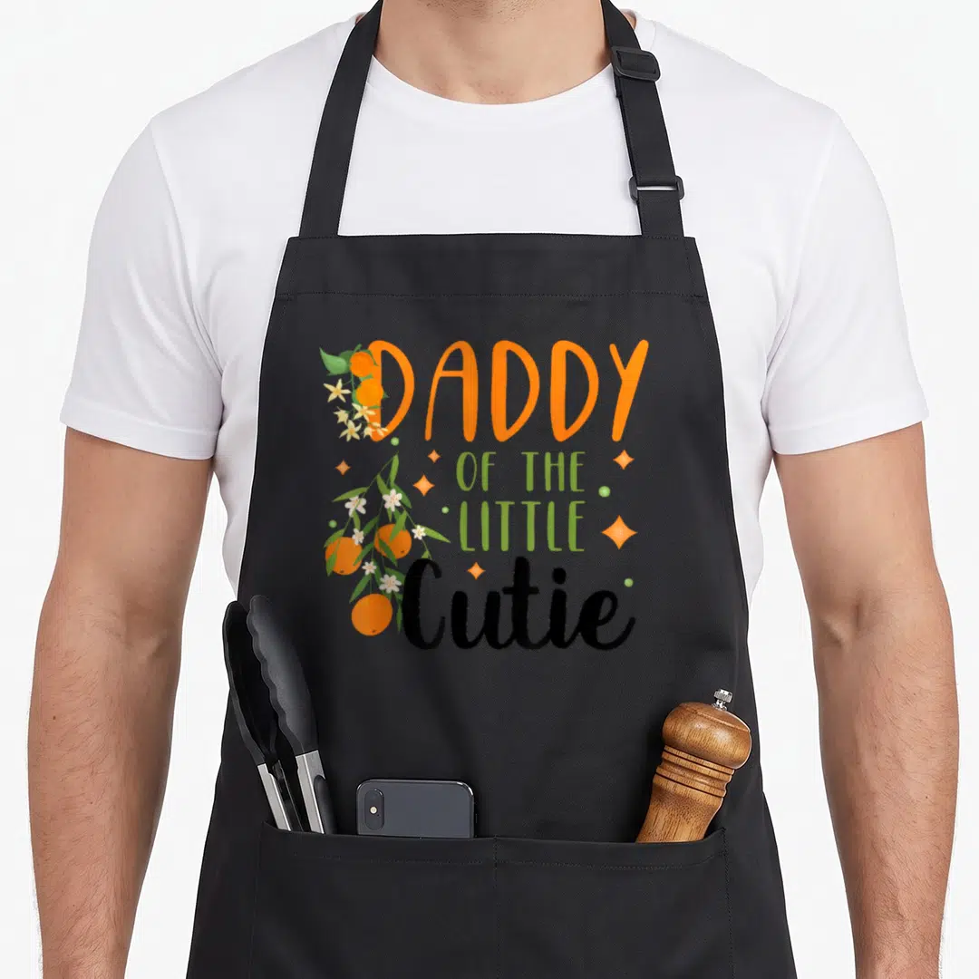 Cutie daddy apron - Daddy of the Little Cutie text with clementines and sparkles — shown on a black cotton canvas apron against a white background