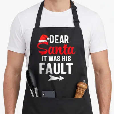 Couple christmas santa apron - Stacked text DEAR Santa IT WAS HIS FAULT with red hat and white arrow pointing right — shown on a black cotton canvas apron against a white background