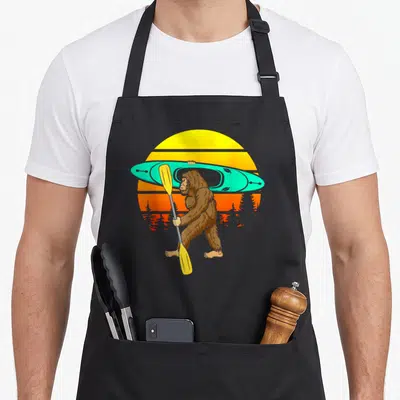 Paddling bigfoot apron - Cartoon Bigfoot carrying turquoise kayak and yellow paddle in front of yellow orange sun — shown on a black cotton canvas apron against a white background