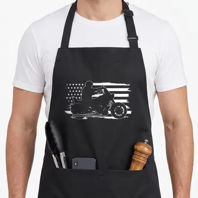 Motorcycle patriotic aprons - White silhouette of a motorcyclist on a distressed American flag pattern — shown on a black cotton canvas apron against a white background
