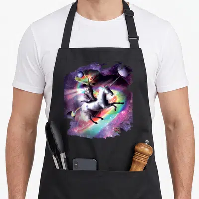 Riding space cat apron - Tabby cat riding leaping white unicorn in purple and pink cosmic space with rainbow and planets — shown on a black cotton canvas apron against a white background