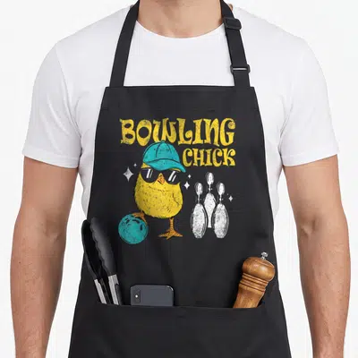 Chick bowl apron - Cartoon yellow chick with blue cap and sunglasses standing on a bowling ball near white pins — shown on a black cotton canvas apron against a white background