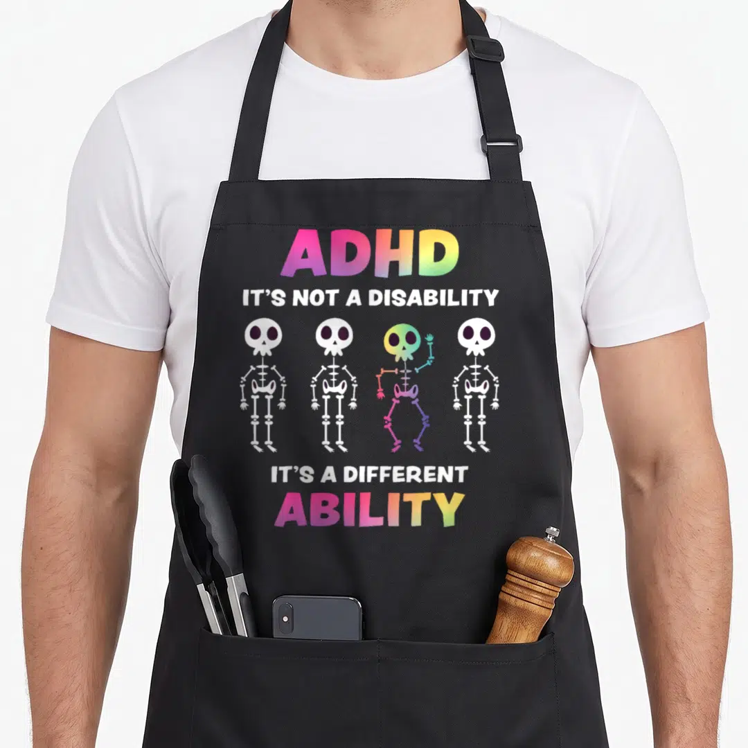 Family skeleton apron - Four cartoon skeletons with rainbow third figure and ADHD awareness text — shown on a black cotton canvas apron against a white background