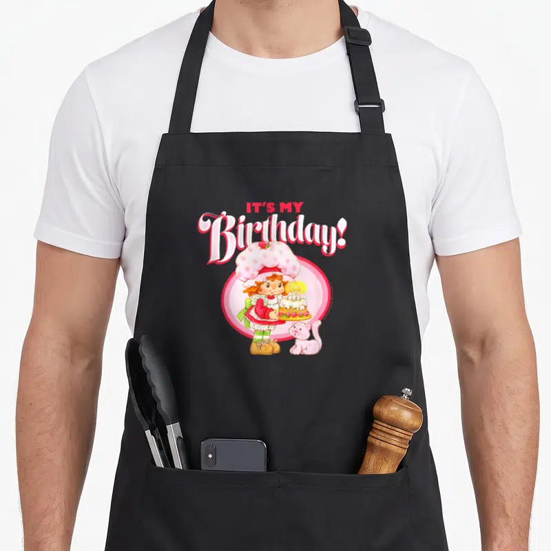 Birthday strawberry apron - Girl in strawberry hat holding cake with pink cat under It's My Birthday text — shown on a black cotton canvas apron against a white background