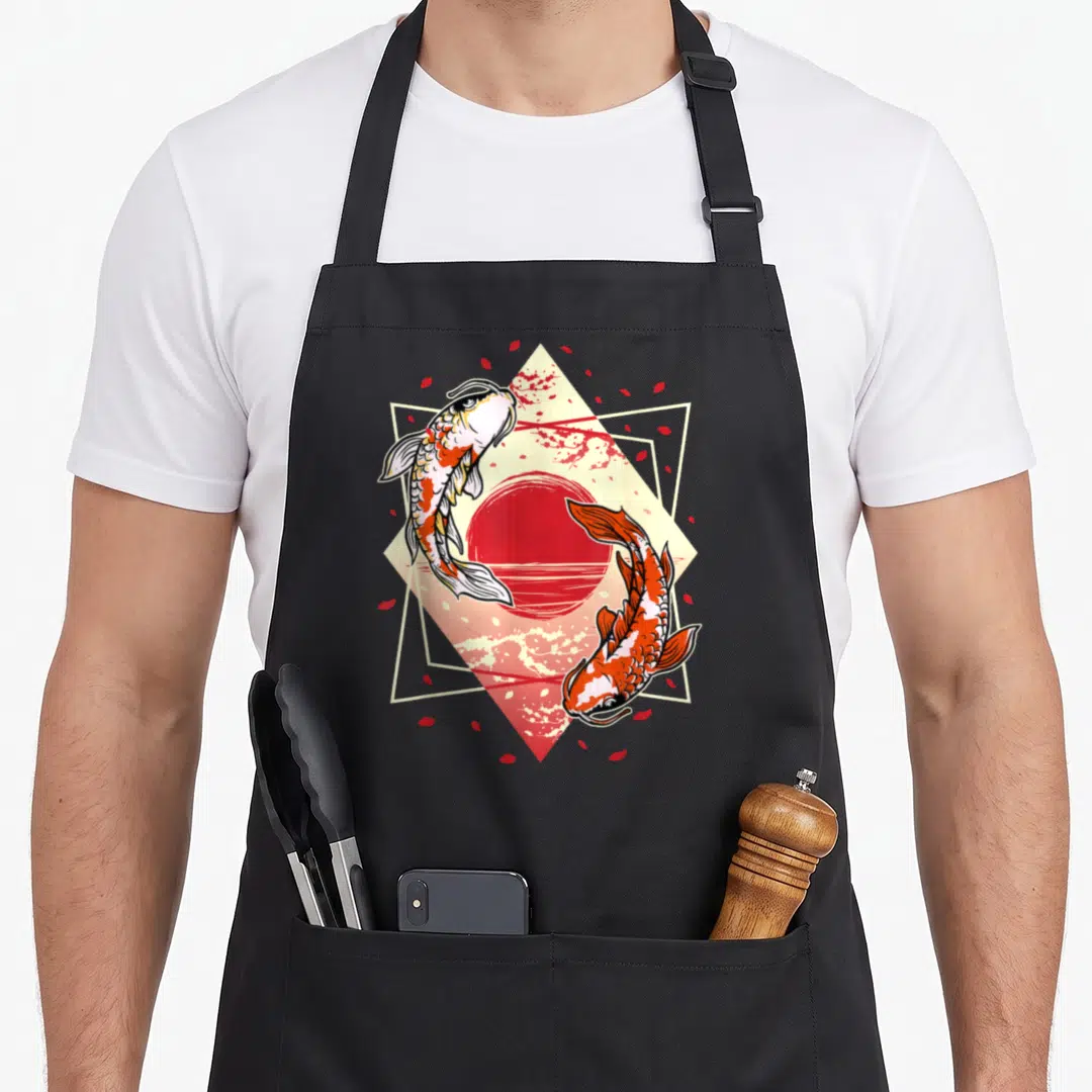 Japanese koi cherry apron - Two koi fish circling red sun in cream diamond frame with red petals — shown on a black cotton canvas apron against a white background