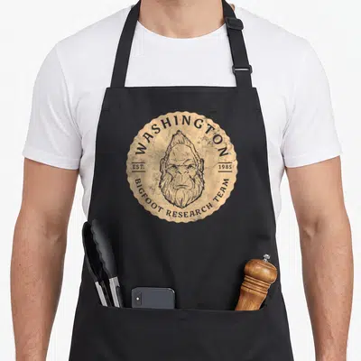 Research washington bigfoot apron - Tan scalloped badge with grey bigfoot head and arched text reading Washington — shown on a black cotton canvas apron against a white background