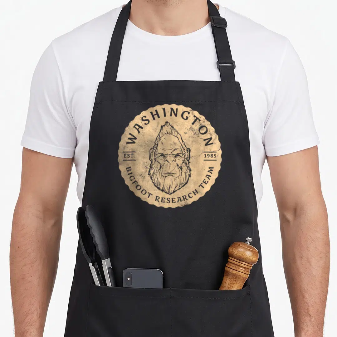 Research washington bigfoot apron - Tan scalloped badge with grey bigfoot head and arched text reading Washington — shown on a black cotton canvas apron against a white background