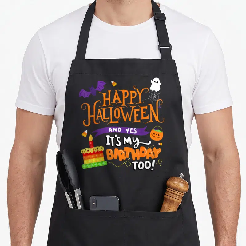 Cute birthday funny halloween apron - Stacked HAPPY HALLOWEEN birthday text with purple bat, white ghost, and rainbow — shown on a black cotton canvas apron against a white background