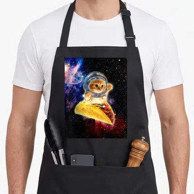 Space black cat aprons - Orange tabby cat in astronaut helmet sitting inside a floating taco against a starry nebula — shown on a black cotton canvas apron against a white background