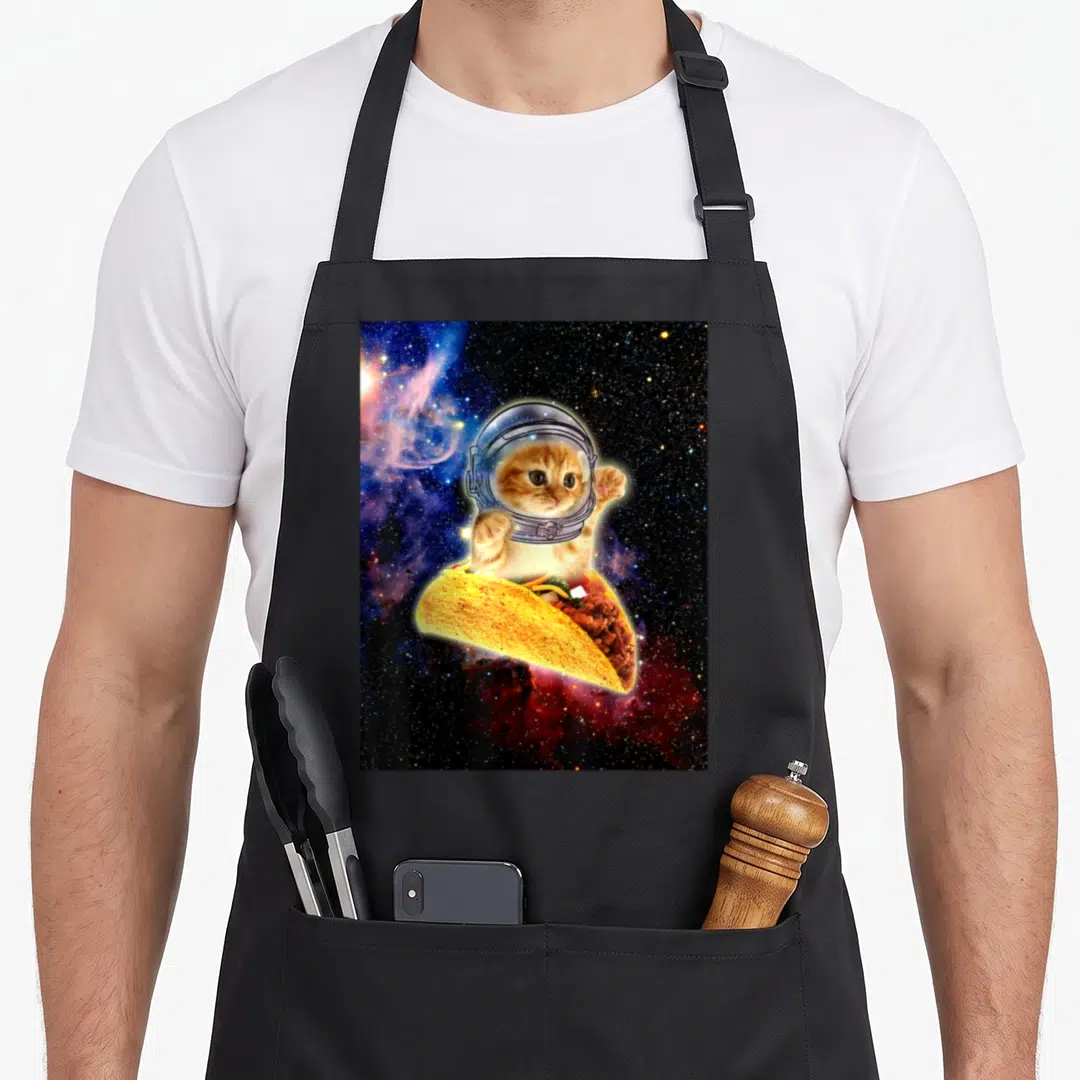 Space black cat aprons - Orange tabby cat in astronaut helmet sitting inside a floating taco against a starry nebula — shown on a black cotton canvas apron against a white background