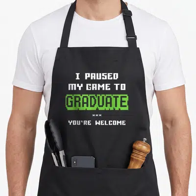 Gamer grade funny apron - I paused my game to graduate you're welcome pixel text white green — shown on a black cotton canvas apron against a white background
