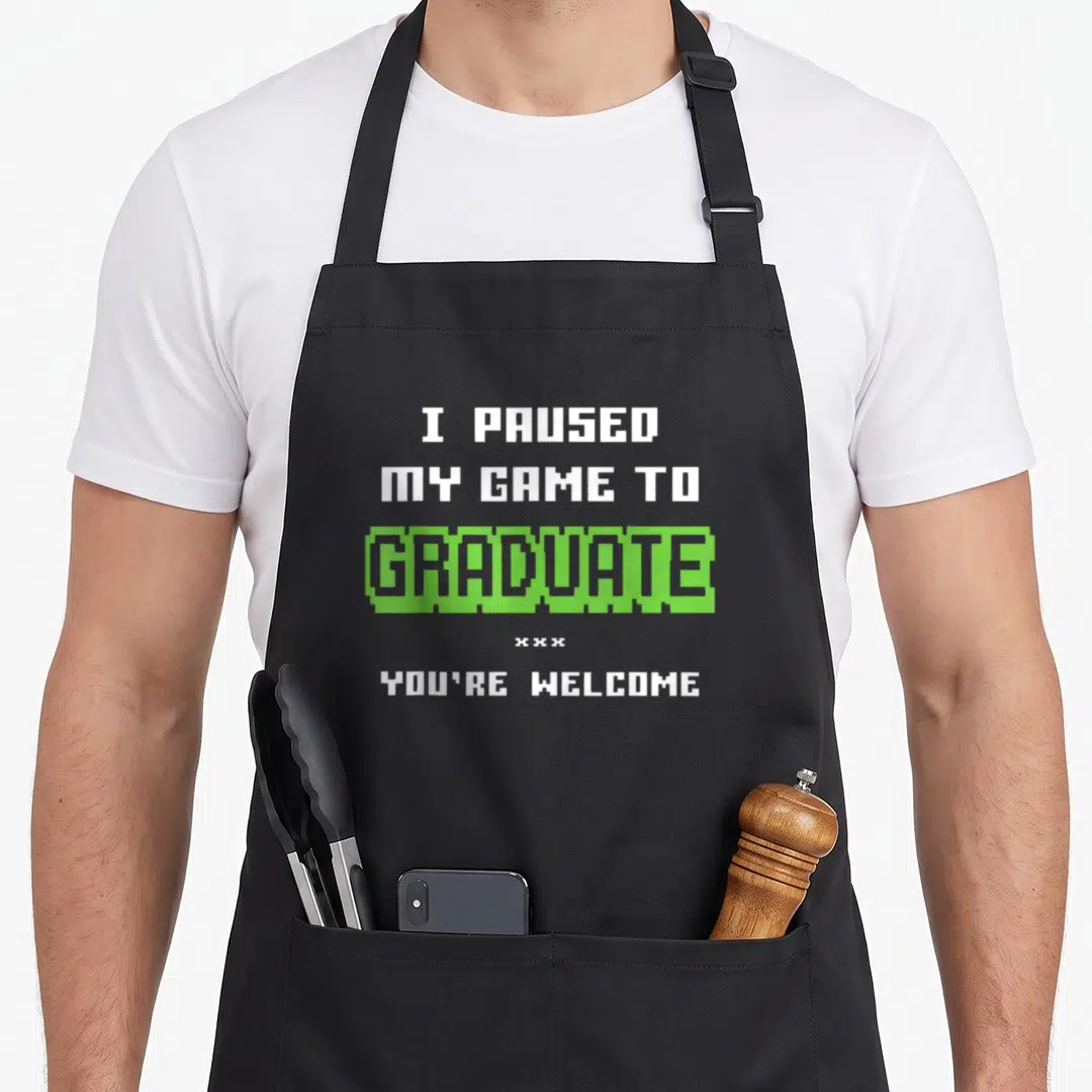 Gamer grade funny apron - I paused my game to graduate you're welcome pixel text white green — shown on a black cotton canvas apron against a white background