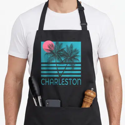 Souvenir charleston apron - Two black palm trees over pink sun and blue stripes with stacked Charleston text — shown on a black cotton canvas apron against a white background