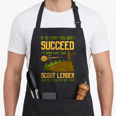 Try scouting america apron - Scout silhouette on mountain with staff and flag, green and yellow text — shown on a black cotton canvas apron against a white background