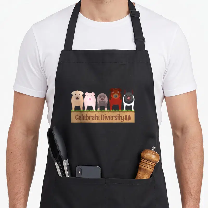 Diversity farm apron - Five cartoon pigs brown pink grey on grass above brown sign with Celebrate Diversity text — shown on a black cotton canvas apron against a white background