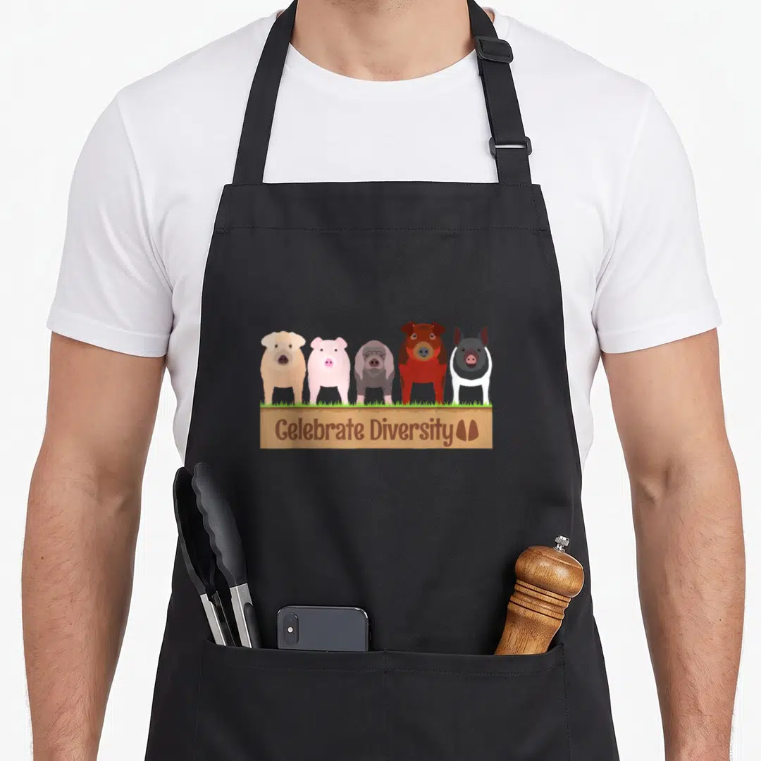 Diversity farm apron - Five cartoon pigs brown pink grey on grass above brown sign with Celebrate Diversity text — shown on a black cotton canvas apron against a white background