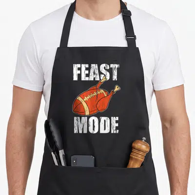 Mode thanksgiving apron - Cartoon football turkey with Feast Mode text in orange and white — shown on a black cotton canvas apron against a white background