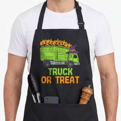 Baby truck apron - Green garbage truck with orange pumpkins and purple witch hat, TRUCK OR TREAT text — shown on a black cotton canvas apron against a white background