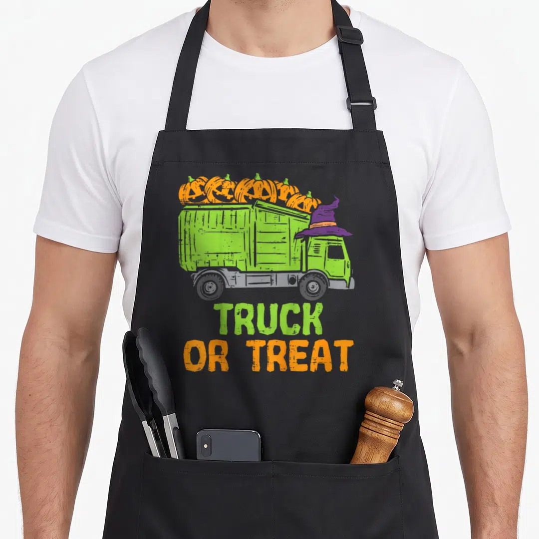 Baby truck apron - Green garbage truck with orange pumpkins and purple witch hat, TRUCK OR TREAT text — shown on a black cotton canvas apron against a white background