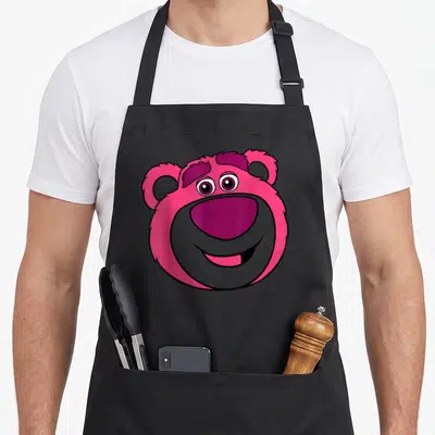 Lotso bear dog breed aprons - Cartoon bear head in pink purple and grey with a smile — shown on a black cotton canvas apron against a white background