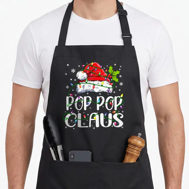 Pop christmas lights apron - Santa hat wrapped in multicolored lights with POP POP CLAUS text and holly — shown on a black cotton canvas apron against a white background