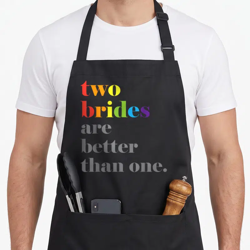 Wedding couple apron - Rainbow gradient text two brides are better than one on grey background — shown on a black cotton canvas apron against a white background