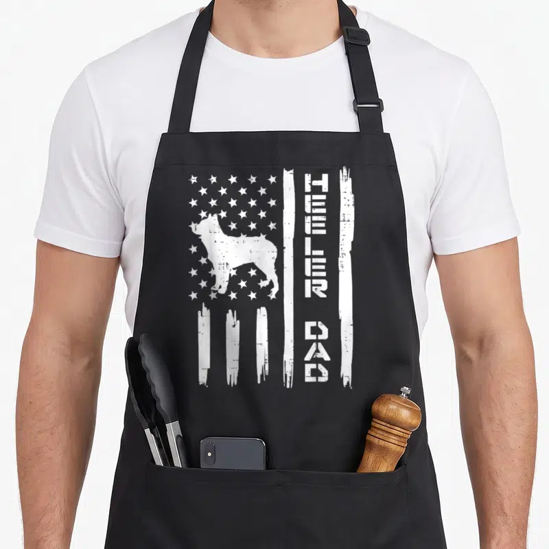 Patriot dog apron - White distressed flag with cattle dog silhouette and stacked heeler dad text on black fabric — shown on a black cotton canvas apron against a white background