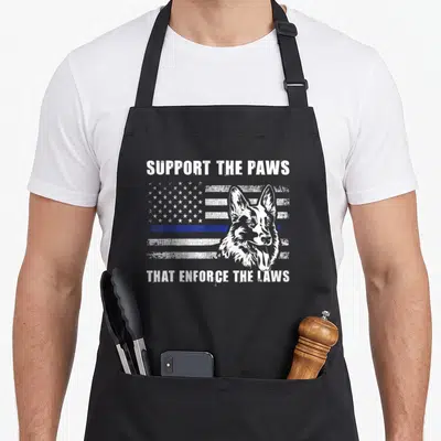 Support line apron - Distressed white dog head over blue line flag with SUPPORT THE PAWS THAT ENFORCE THE LAWS text — shown on a black cotton canvas apron against a white background