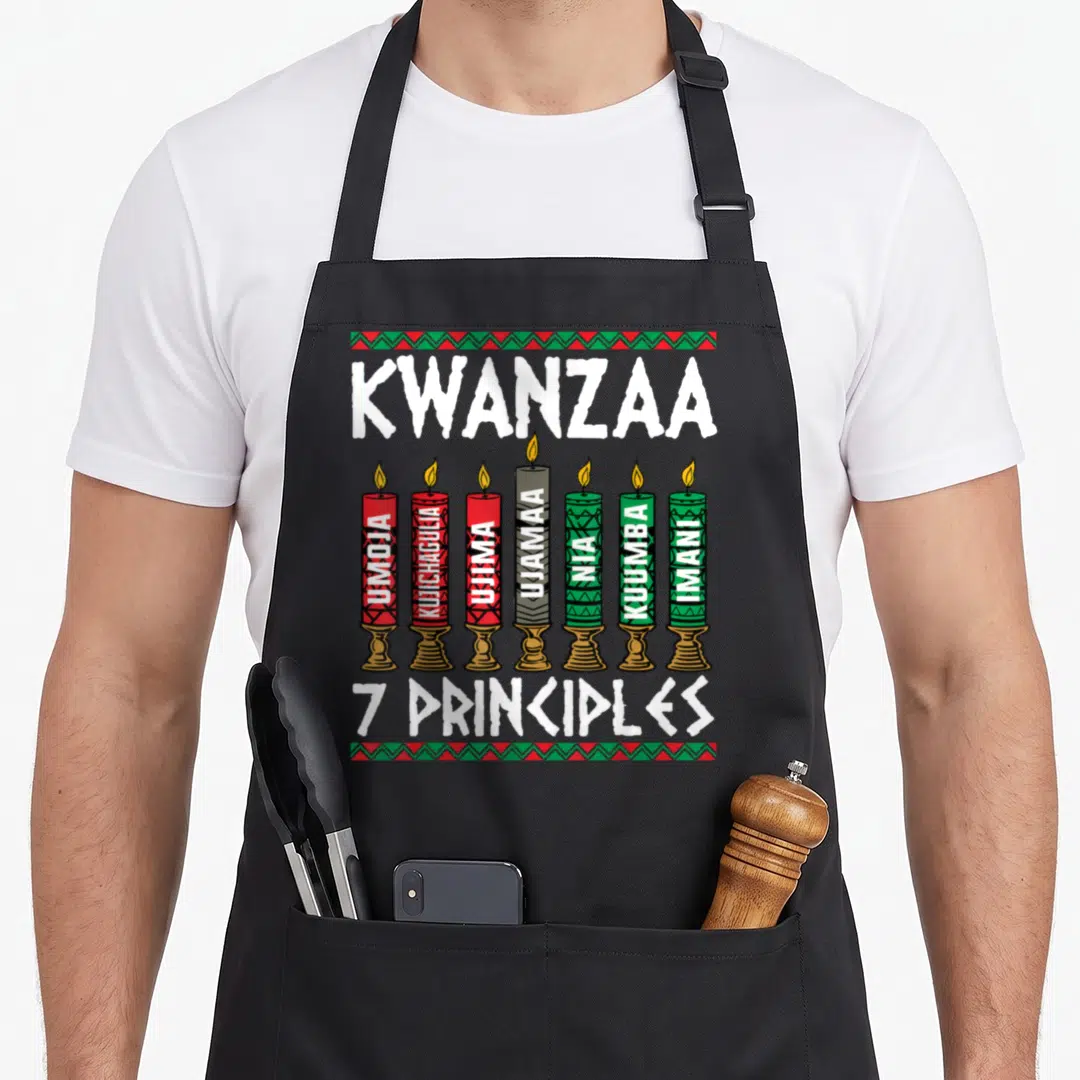 Kwanzaa production quotes apron - Seven candles labeled Umoja Imani with decorative borders in red green black — shown on a black cotton canvas apron against a white background