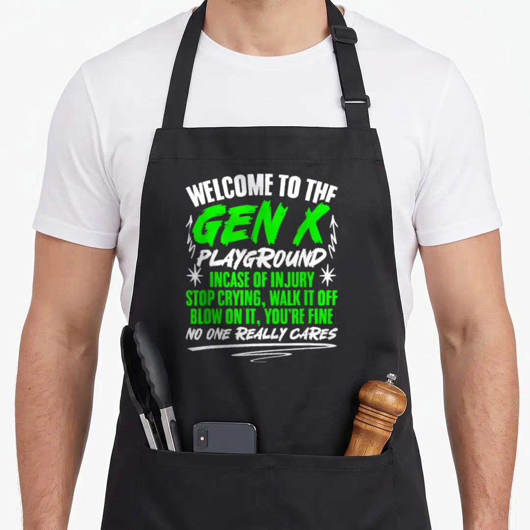 Welcome gen x apron - Green and white stacked text reading Welcome to the Gen X Playground with arrows and asterisks — shown on a black cotton canvas apron against a white background