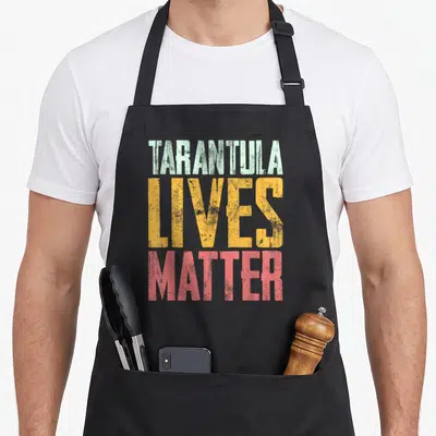 Live tarantula apron - Stacked distressed text reading TARANTULA LIVES MATTER in green, yellow, and red block letters — shown on a black cotton canvas apron against a white background