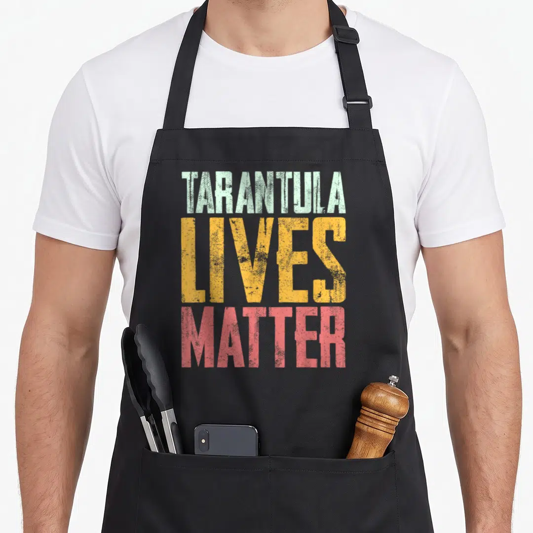 Live tarantula apron - Stacked distressed text reading TARANTULA LIVES MATTER in green, yellow, and red block letters — shown on a black cotton canvas apron against a white background